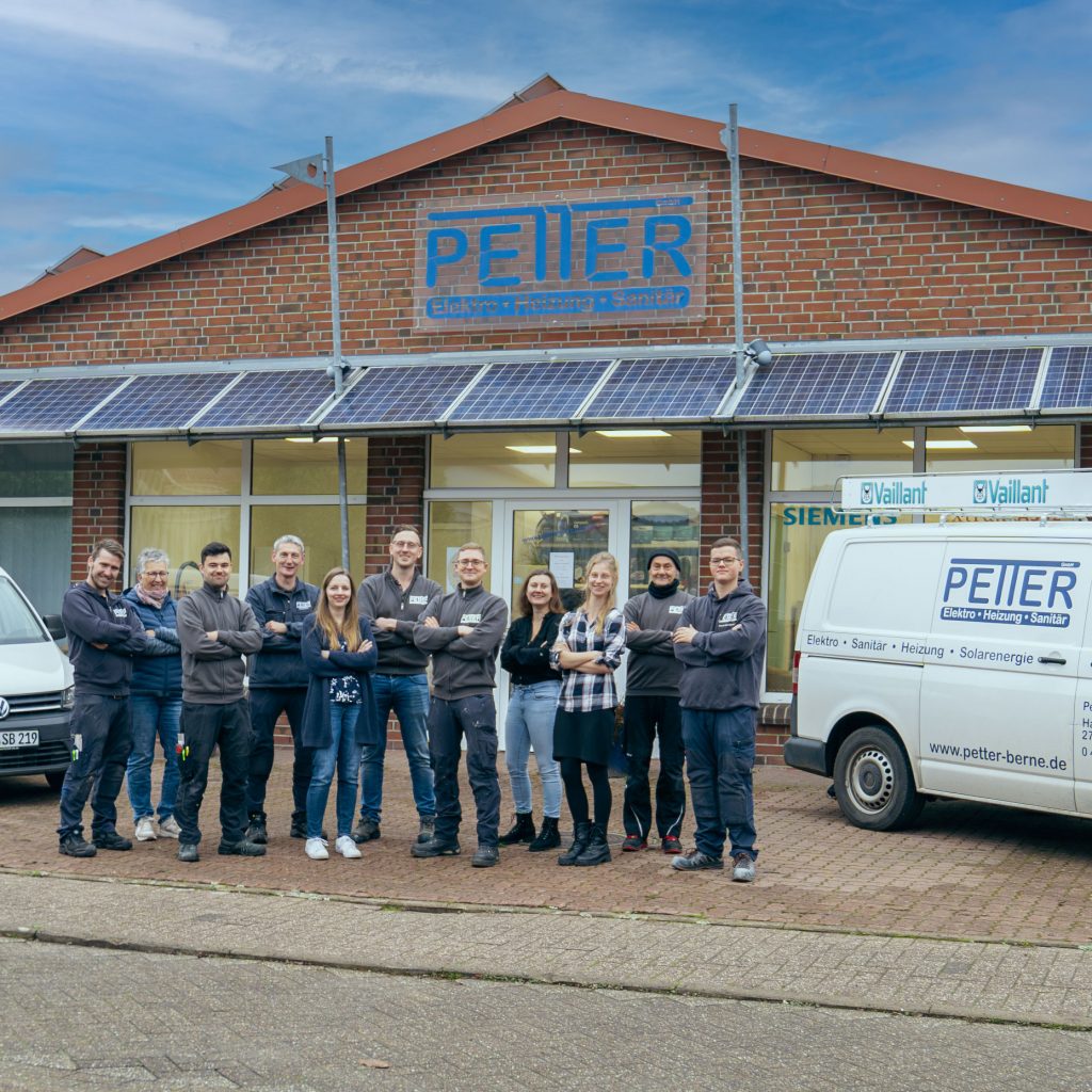 Ein Team von Mitarbeitenden in Arbeitskleidung steht mit verschränkten Armen vor einem Backsteingebäude mit Solarpaneelen und großem blauem Firmenlogo »PETTER«, rechts davon ein beschrifteter Firmenwagen.